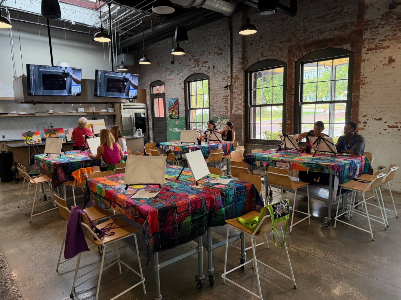 An art class in a bright, industrial-style studio with large windows, tables covered with colorful patchwork tablecloths, and easels with blank canvases. There are a few students seated at the tables painting, with two women working together near the