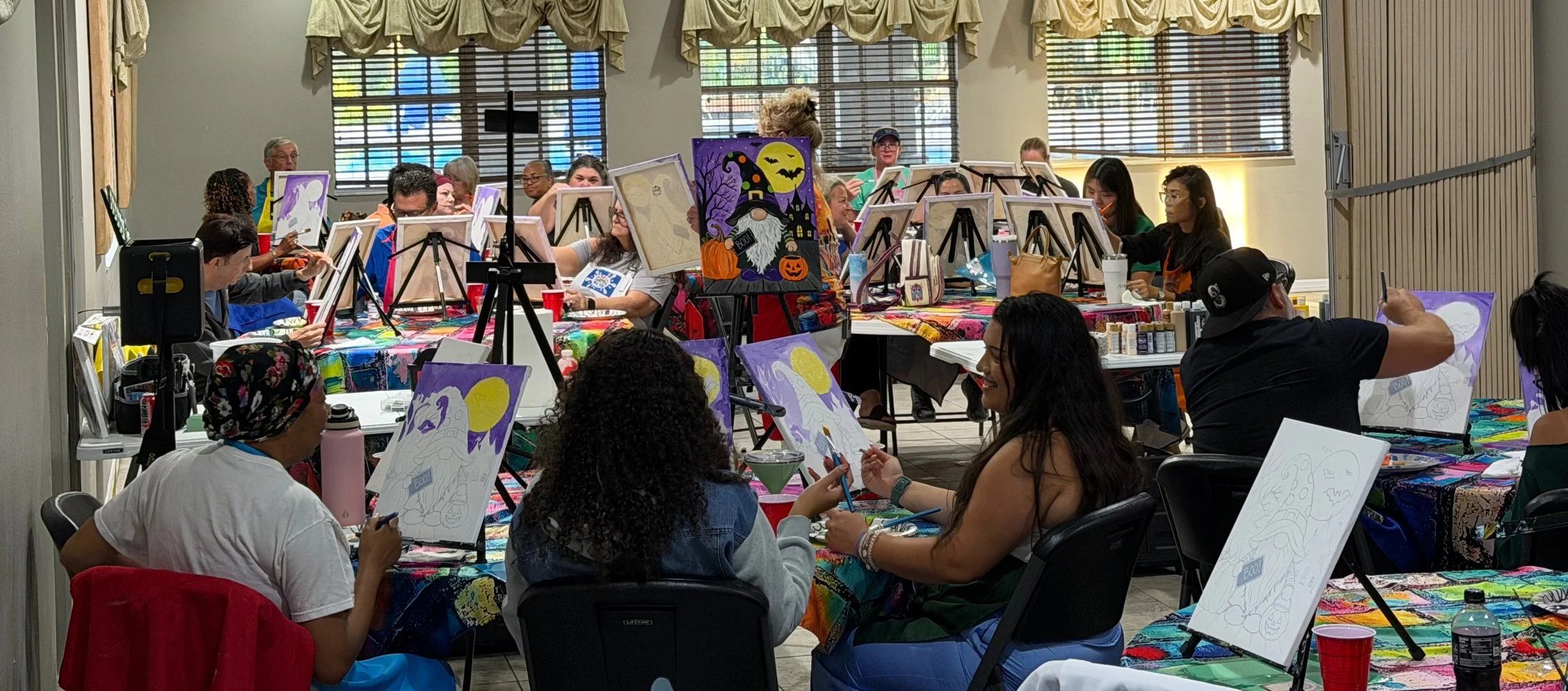 People participating in a Halloween-themed painting class, painting spooky witch and pumpkin scenes on canvases, with autumn decorations and colorful tablecloths.