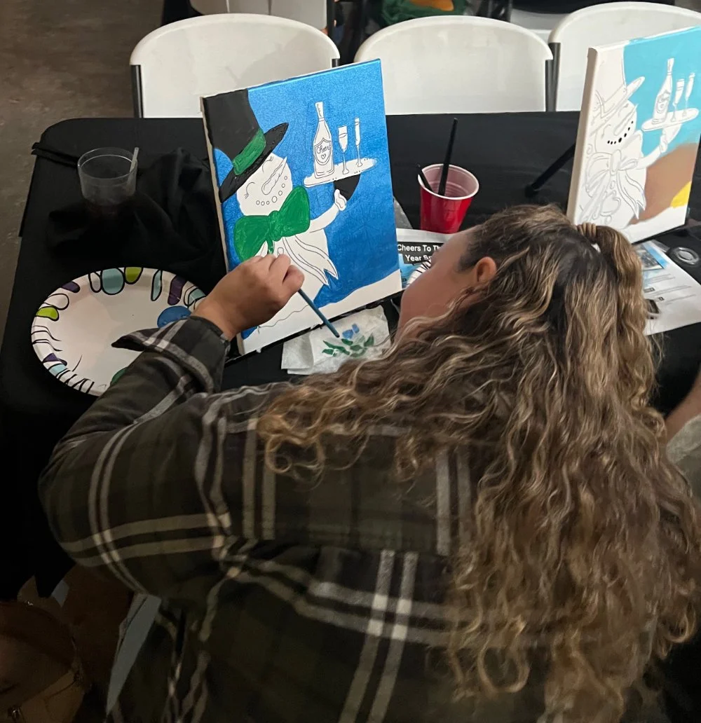 A woman with curly hair painting a picture of a cartoon character of a unicorn with a green bow tie, black hat, and holding a tray with a bottle and two glasses. There are other paintings, brushes, a disposable plate, and cups on the table.