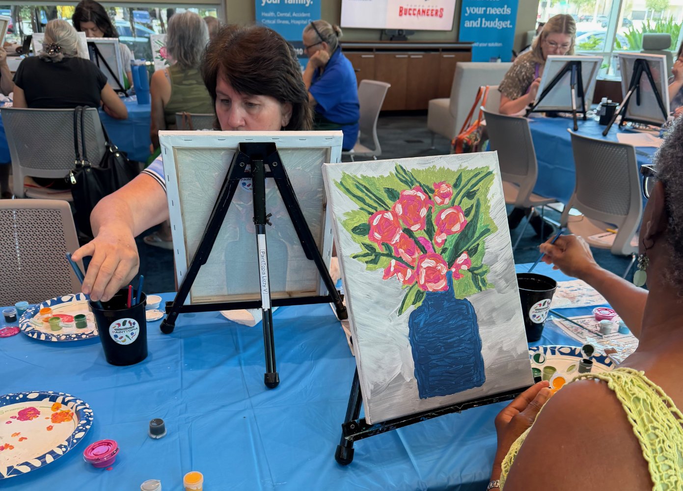 A group of women participating in a painting activity at a restaurant or community center, with a woman painting a colorful floral arrangement in a blue vase on her canvas and others working on their artwork at tables in the background.