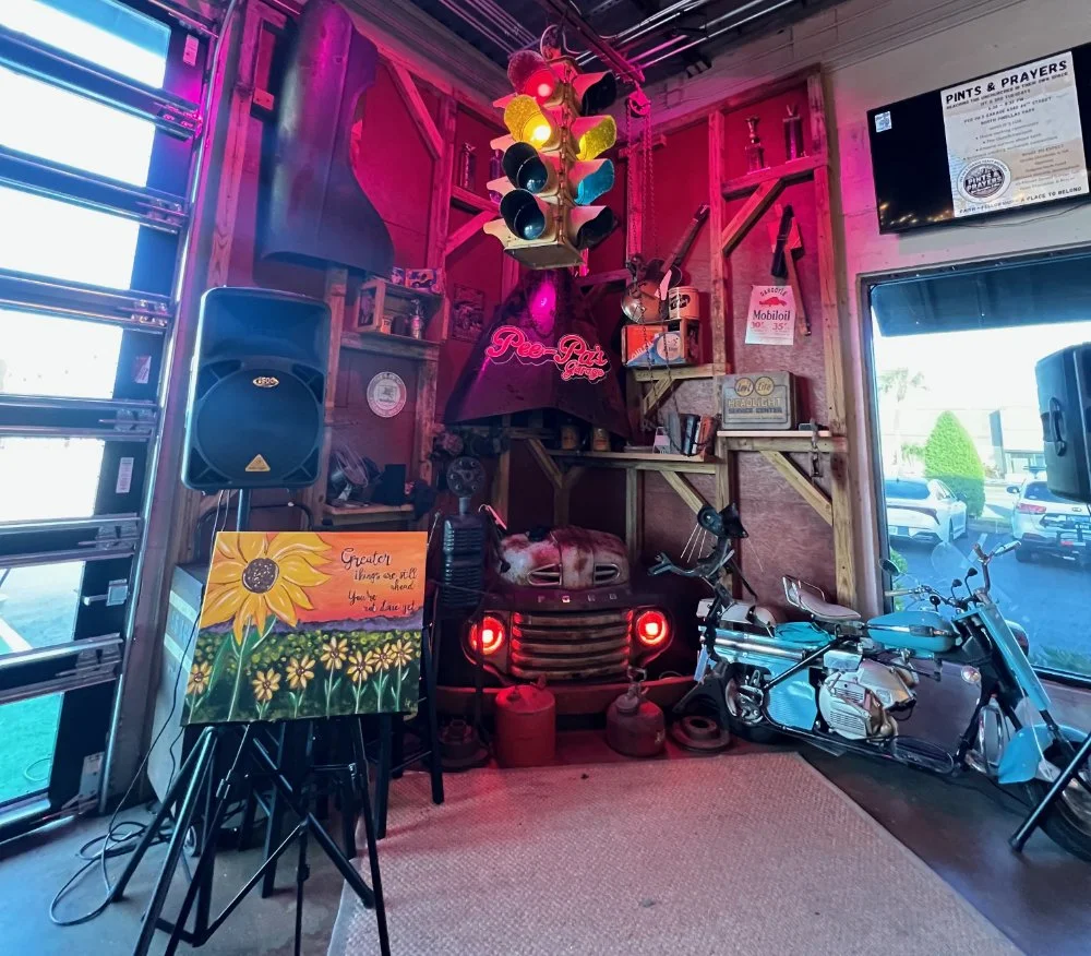 Decorated corner of a vintage-themed establishment with a motorcycle, a toy car, a traffic light, and artwork. Neon sign reads 'Peach State Garage' and there are various signs and collectibles on the wooden walls.