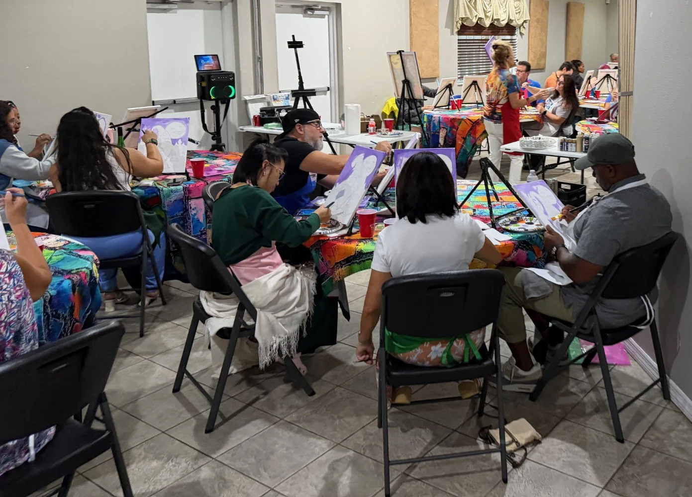 People participating in a group painting class indoors, with each person working on a painting of a full moon on canvas. The room is decorated with colorful tablecloths and painting supplies.