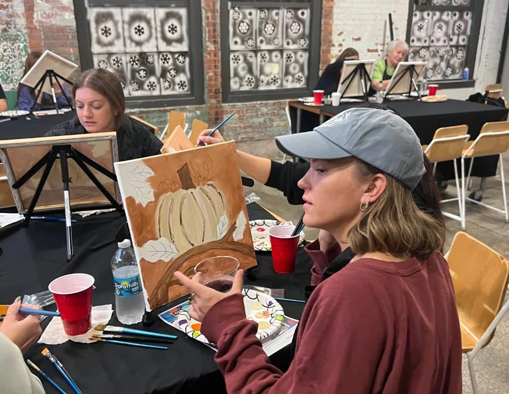 Woman with light brown hair wearing a gray cap and maroon hoodie painting an autumn-themed picture of a pumpkin with leaves on a canvas at an art class.