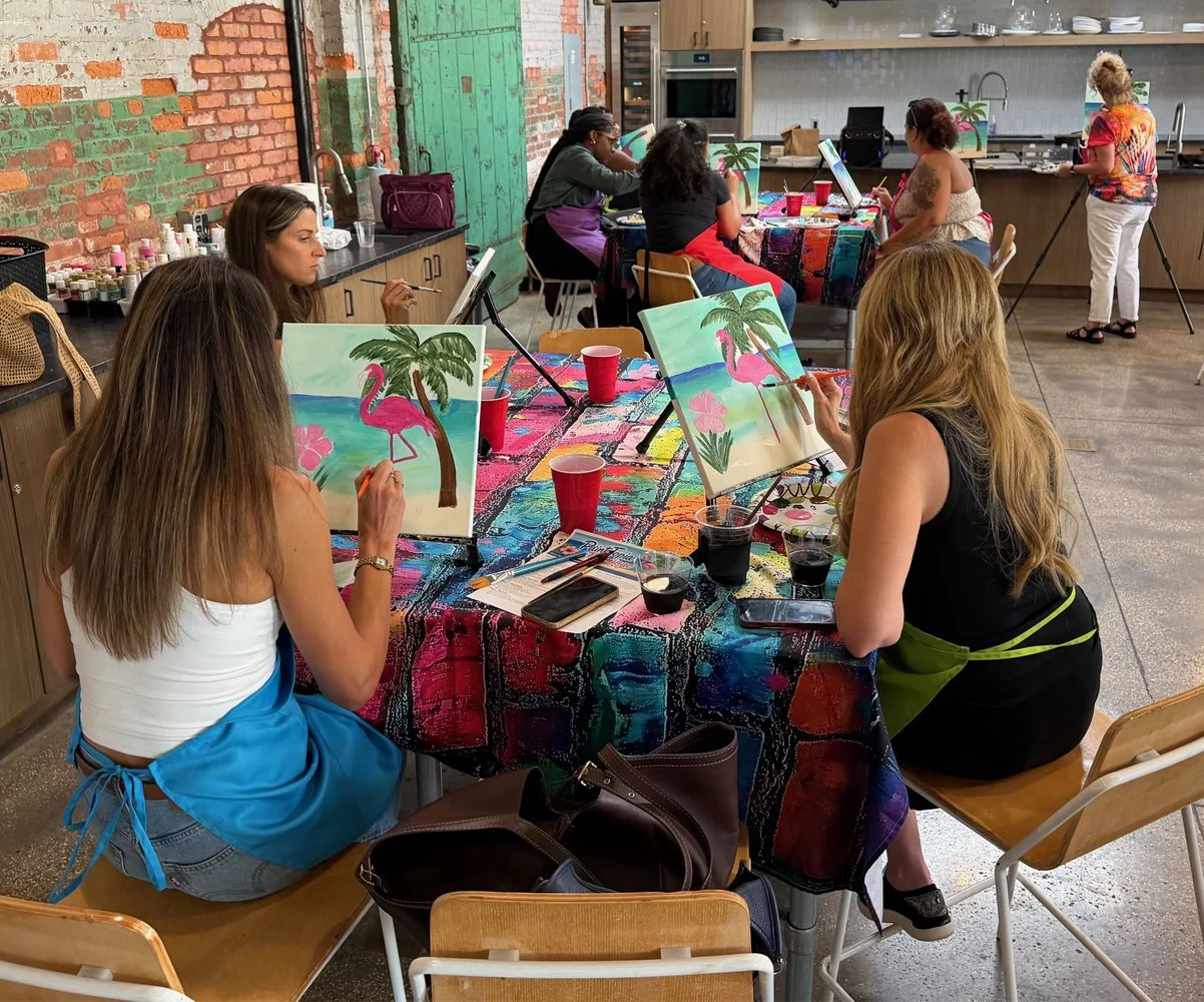 People participating in a group painting class, creating tropical beach scenes with pink flamingos, green palm trees, and a colorful tablecloth in an art studio with exposed brick walls.