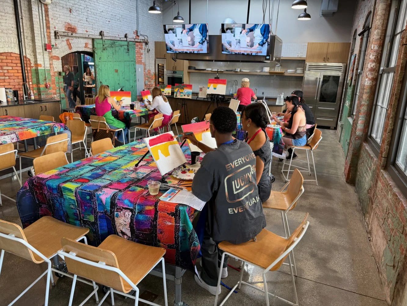 People participating in a painting class in an artistic, industrial-style room with colorful tables, brick walls, and large windows.