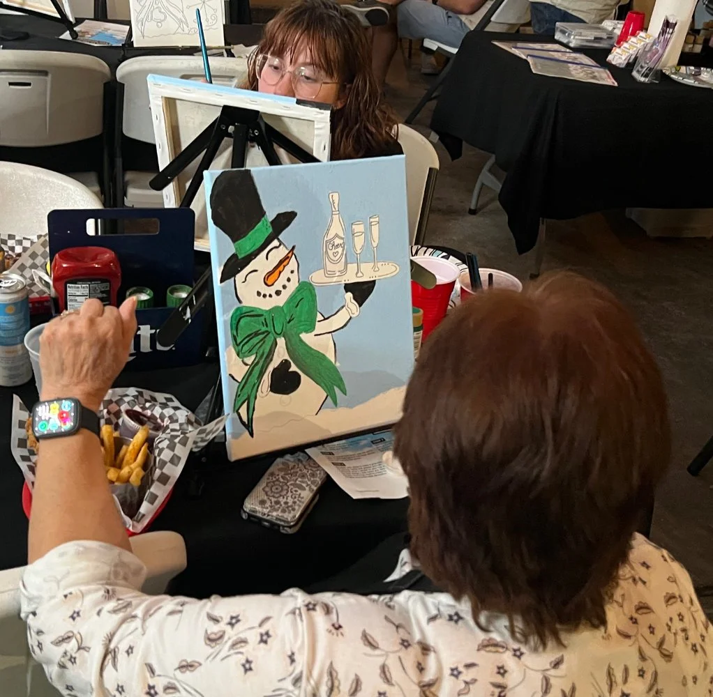 A woman painting a snowman with a hat, green scarf, and carrot nose on a canvas at an art event. The snowman is holding a tray with a bottle and glasses. The scene includes art supplies, snacks, and participants.