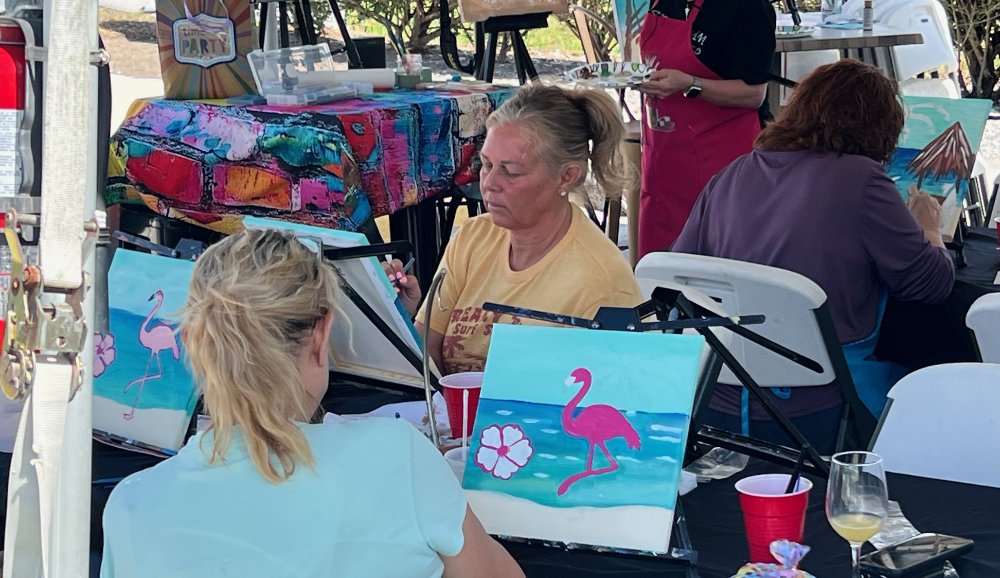 People painting beach scenes with pink flamingos on small canvases at an outdoor event table.