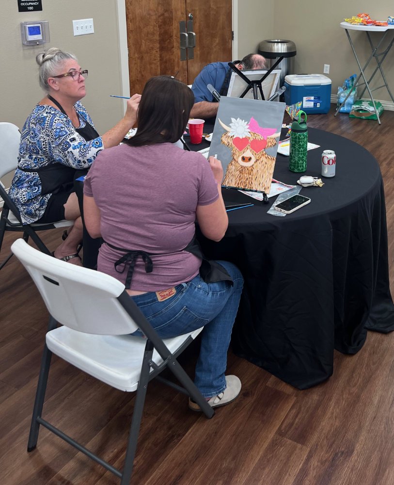 Three people sitting at a black-covered round table, engaged in art or craft activities. One person with blonde hair and glasses is seated on the left, another with dark hair in a purple shirt is in the middle, and a person with a bald head is on the