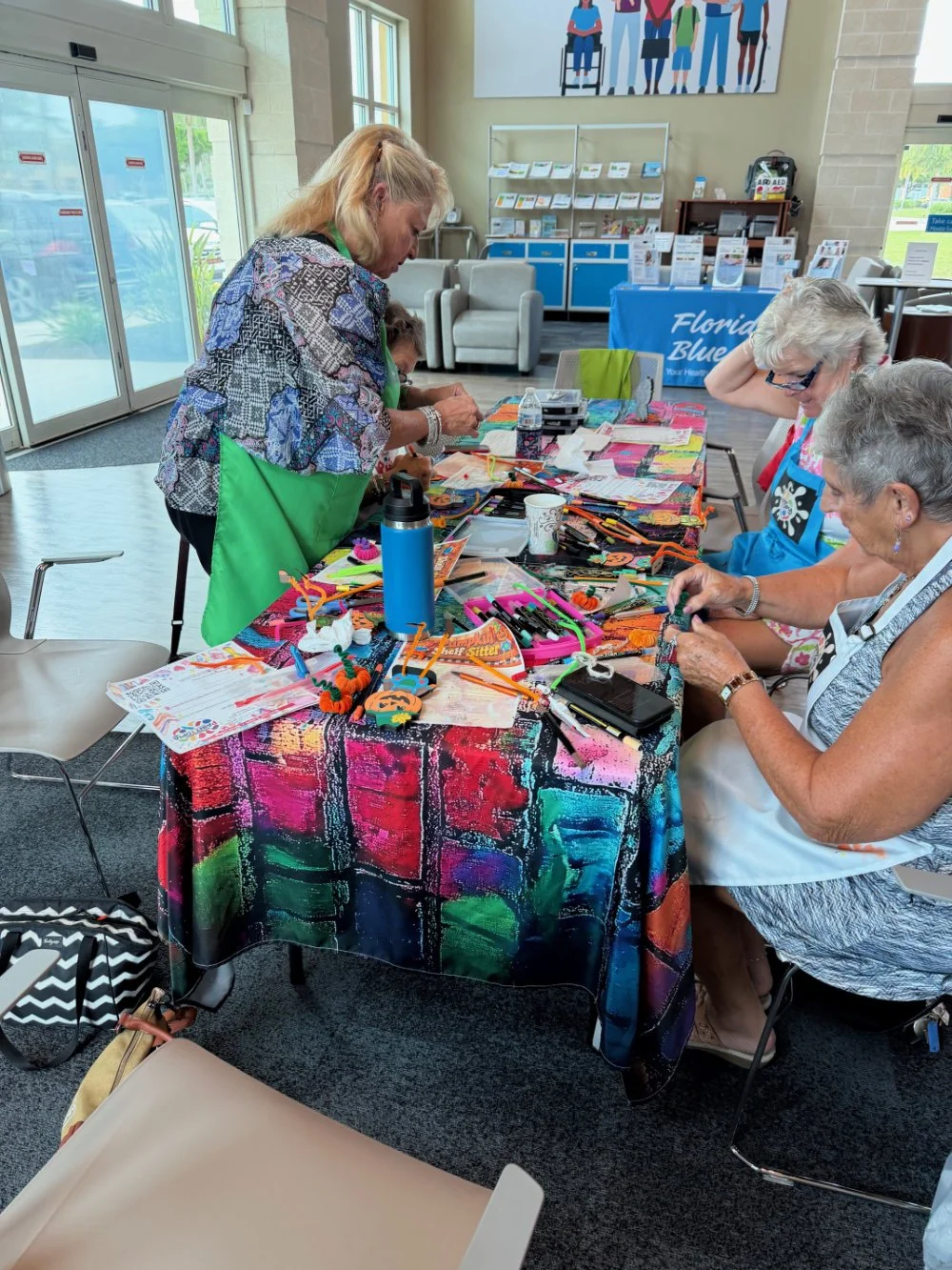 Women participating in a craft workshop at a colorful table inside a community center, with craft supplies and completed projects visible.