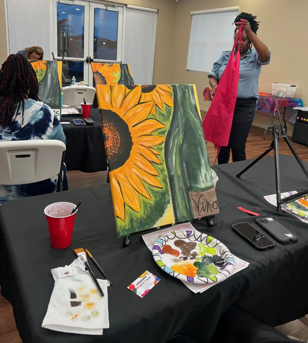 An art class with women painting sunflowers on canvases. One woman is preparing her materials, while others are focused on their paintings. The room has large windows, and art supplies are scattered on the tables.