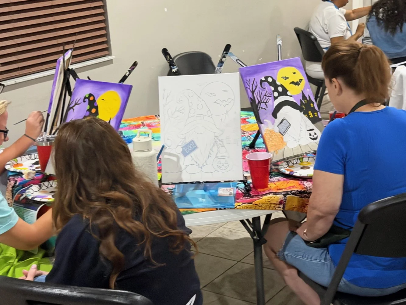 People painting Halloween-themed pictures at a table with colorful tablecloth and red cups.