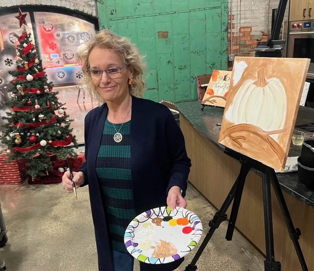 A woman with curly blonde hair, glasses, and a dark blazer standing next to an easel with a pumpkin painting. She is holding a paper plate with paint and a paintbrush. Christmas decorations, including a Christmas tree with ornaments and a red ribbon,