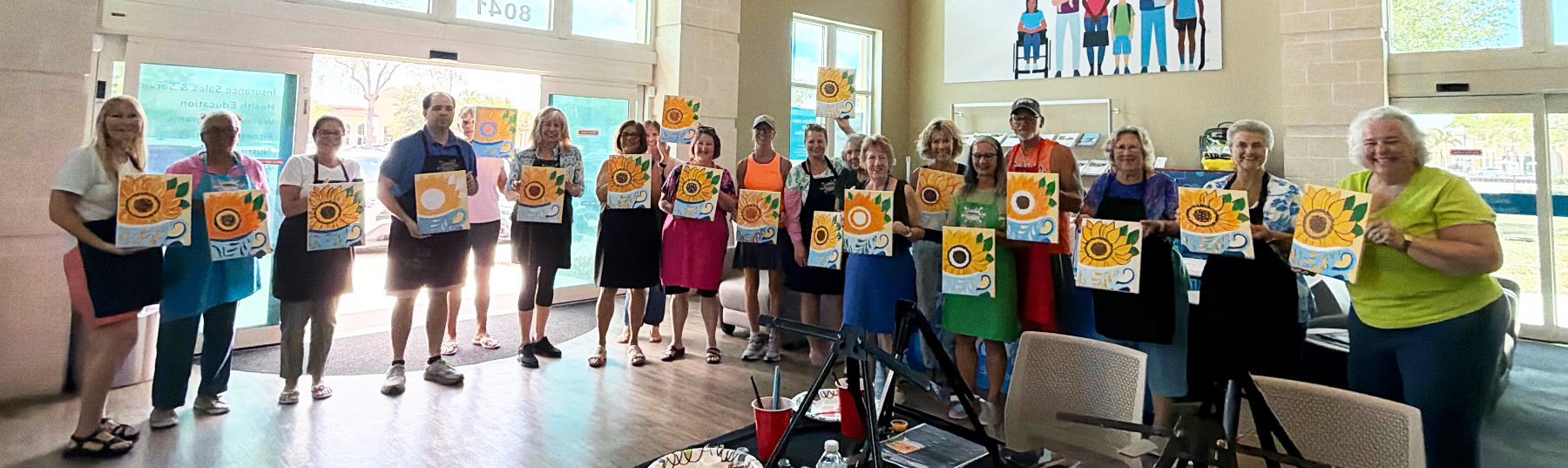 A group of people standing inside a room, each holding a canvas painting of a sunflower with various designs and colors. The group appears to be at a painting class or workshop.