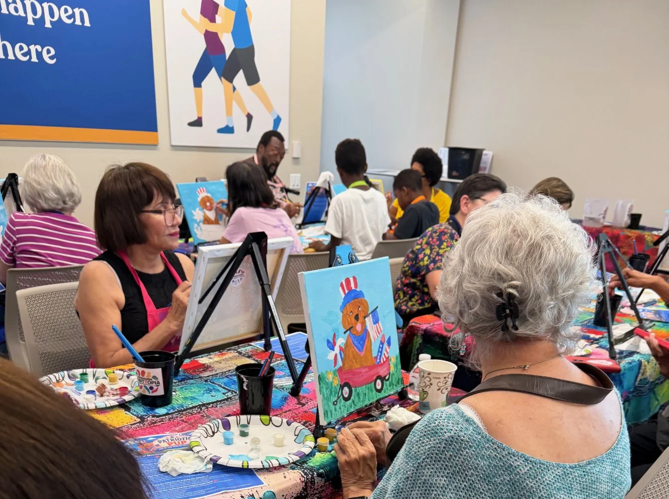 A group of people participating in a painting class, creating patriotic-themed artwork featuring a bear in a red, white, and blue outfit. The setting appears to be a community center or art studio with painting supplies on the tables.