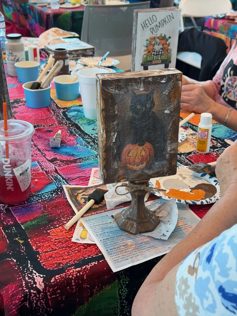 A handcrafted Halloween decoration featuring a black cat with green eyes sitting on a carved pumpkin, painted on a rectangular wooden board, displayed on a table during a craft event.
