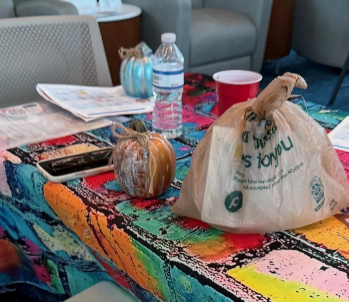 A table covered with a colorful tablecloth contains a small pumpkin, a plastic bag, a water bottle, a smartphone, a red cup, a small ceramic pumpkin, and some papers in a restaurant or cafe setting.