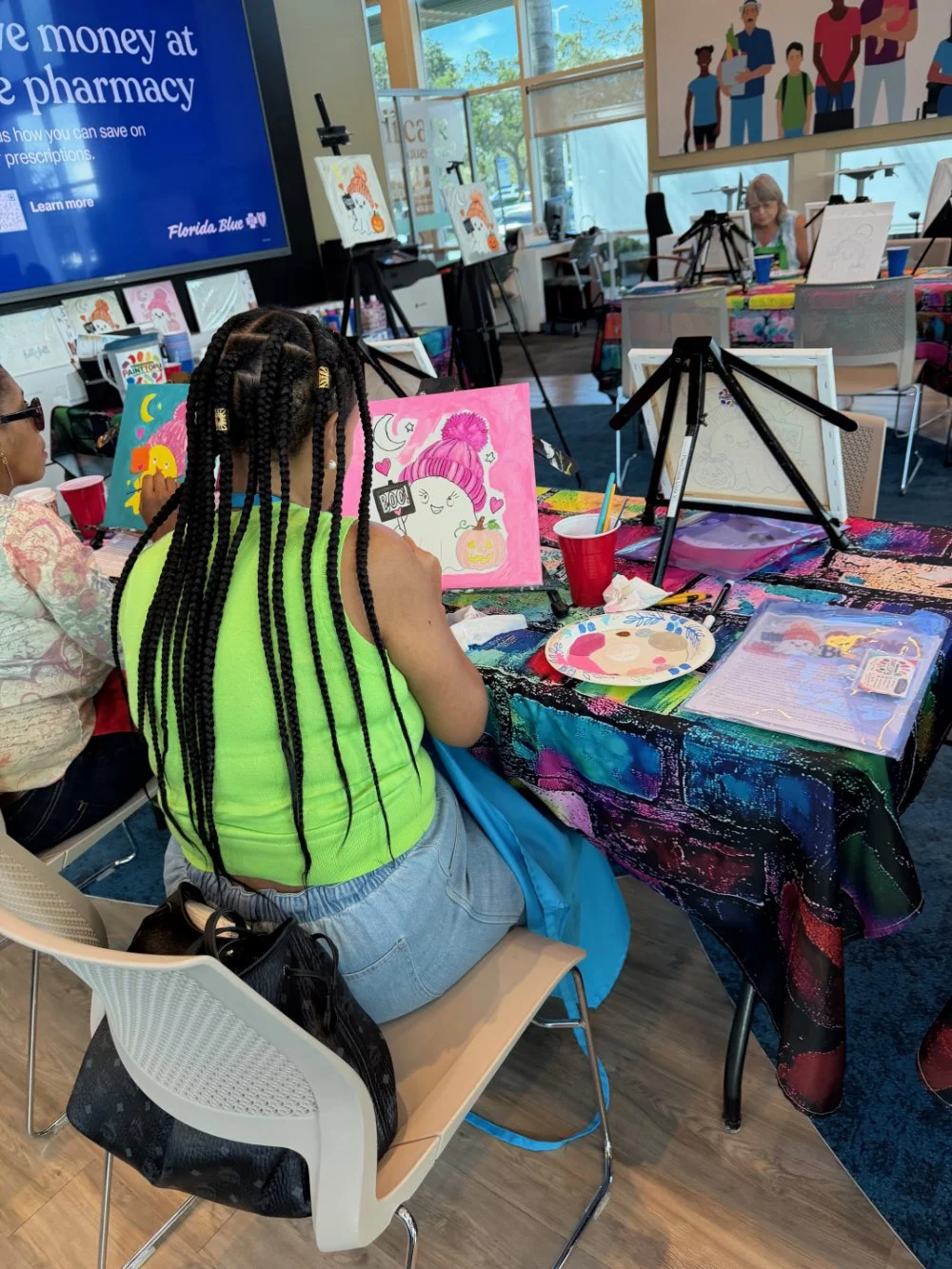 Children participating in a painting class at a café or community center, with colorful artwork and supplies on the table, and other children painting at nearby tables.