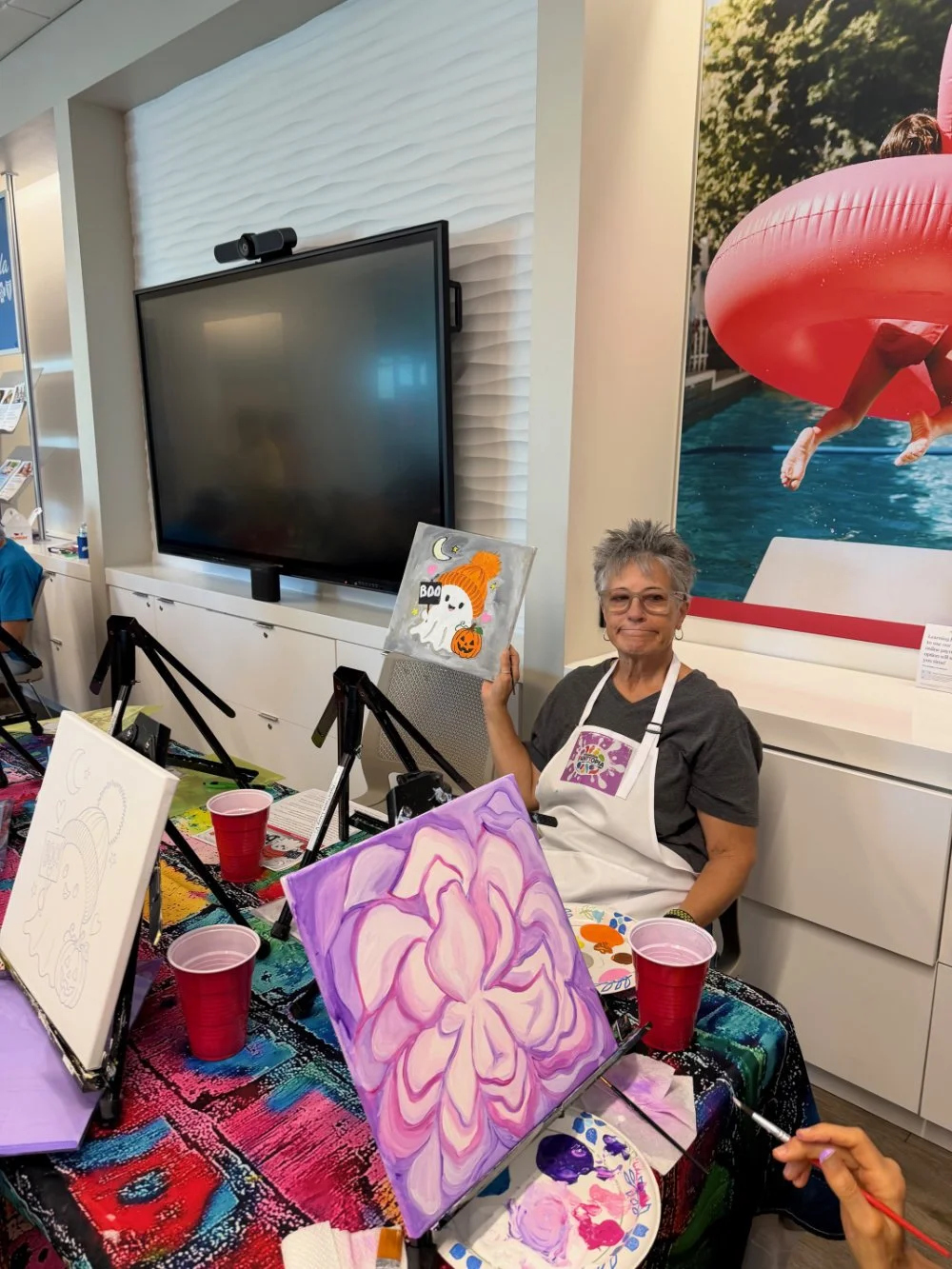 A woman sitting at a table with a colorful tablecloth, holding a Halloween-themed painting of a ghost with a pumpkin and moon. The table has several paintings in progress, red plastic cups, and painting supplies. There is a large flat-screen TV and a