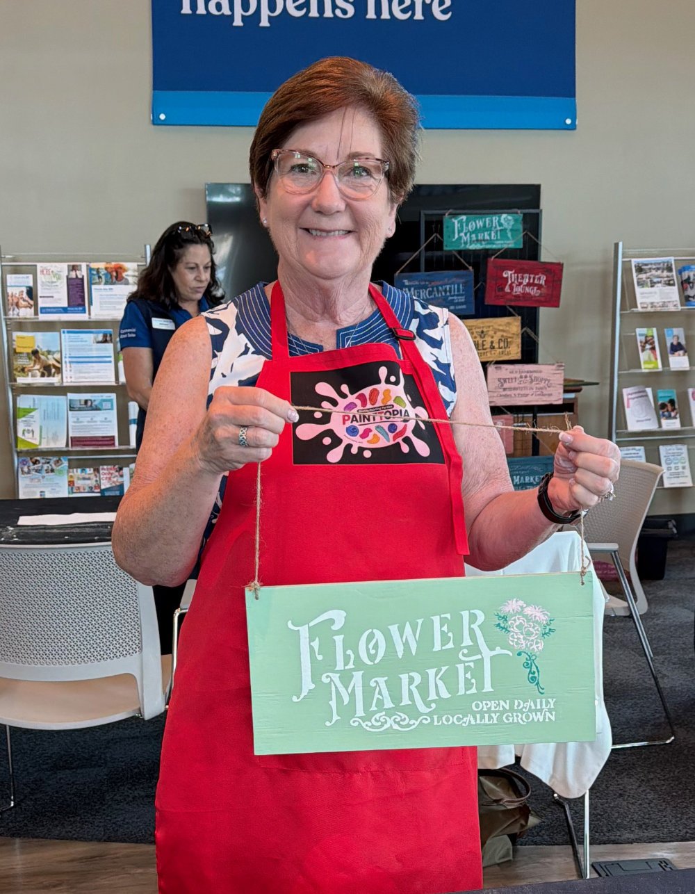 A smiling woman with short brown hair and glasses holding a painted sign that reads, 'Flower Market, Open Daily, Locally Grown,' in an indoor setting with informational displays and another woman in the background.