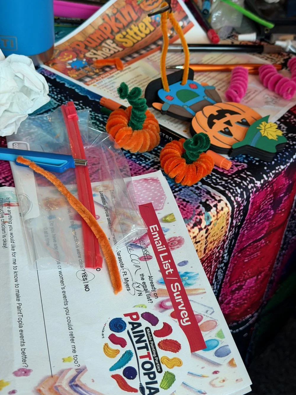 Craft supplies on a table, including pipe cleaners shaped like pumpkins, a pumpkin-themed craft, and various art tools and papers.