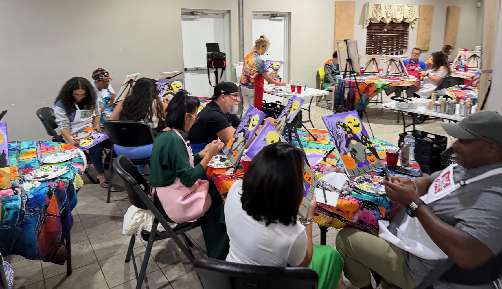 People participating in a Halloween-themed painting class with spooky artwork on canvas, colorful tablecloths, and art supplies in a decorated room.