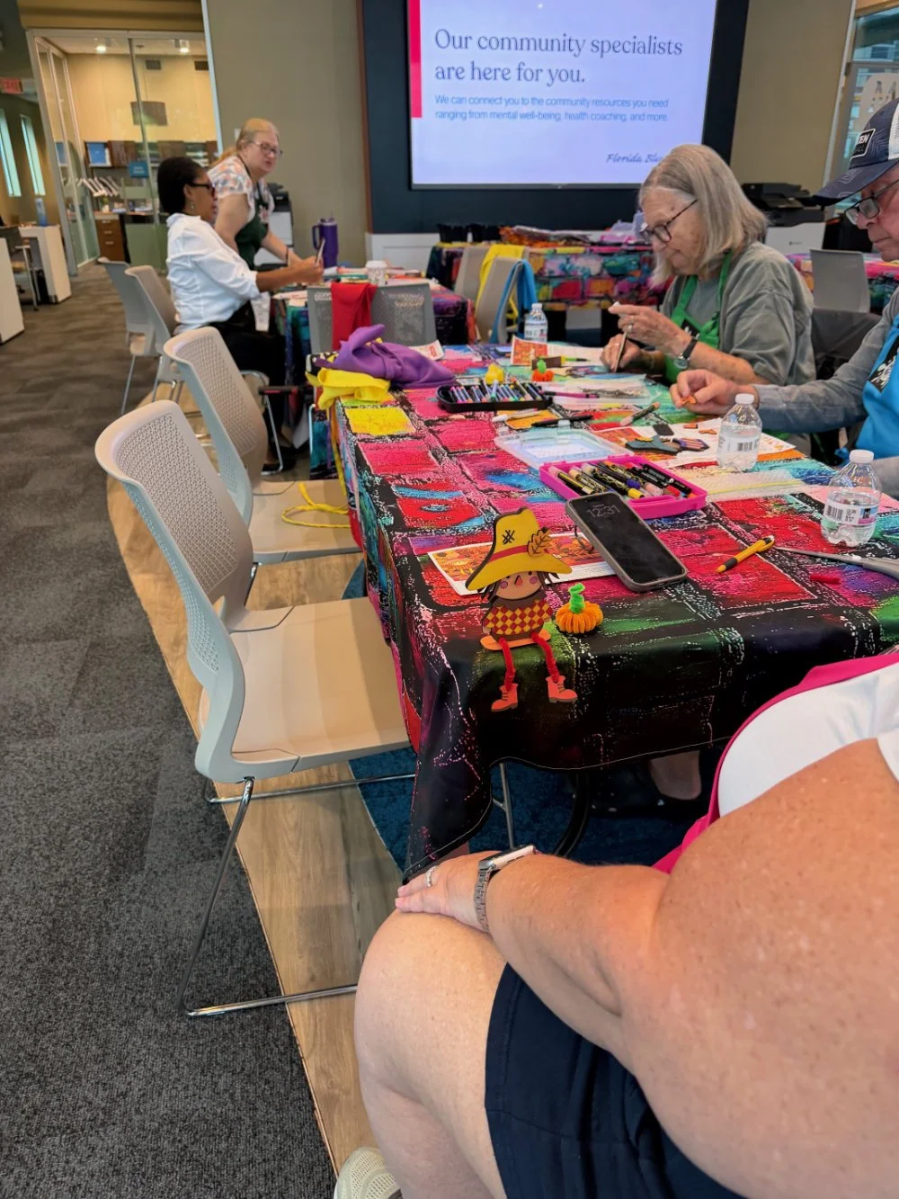 A group of people at a craft workshop with art supplies on a colorful table and a screen displaying information about community specialists.