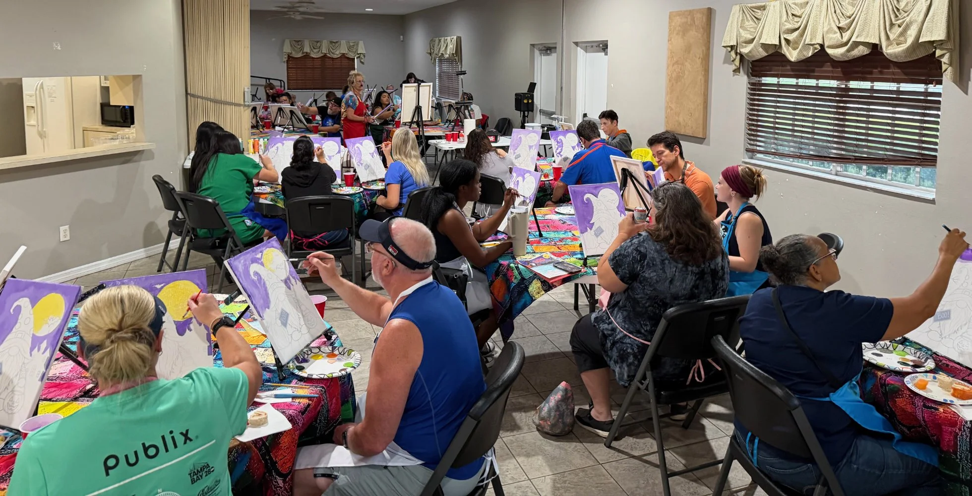 A diverse group of people participating in a group painting class, seated around tables with art supplies, in a well-lit room with windows and curtains, supervised by an instructor at the front.