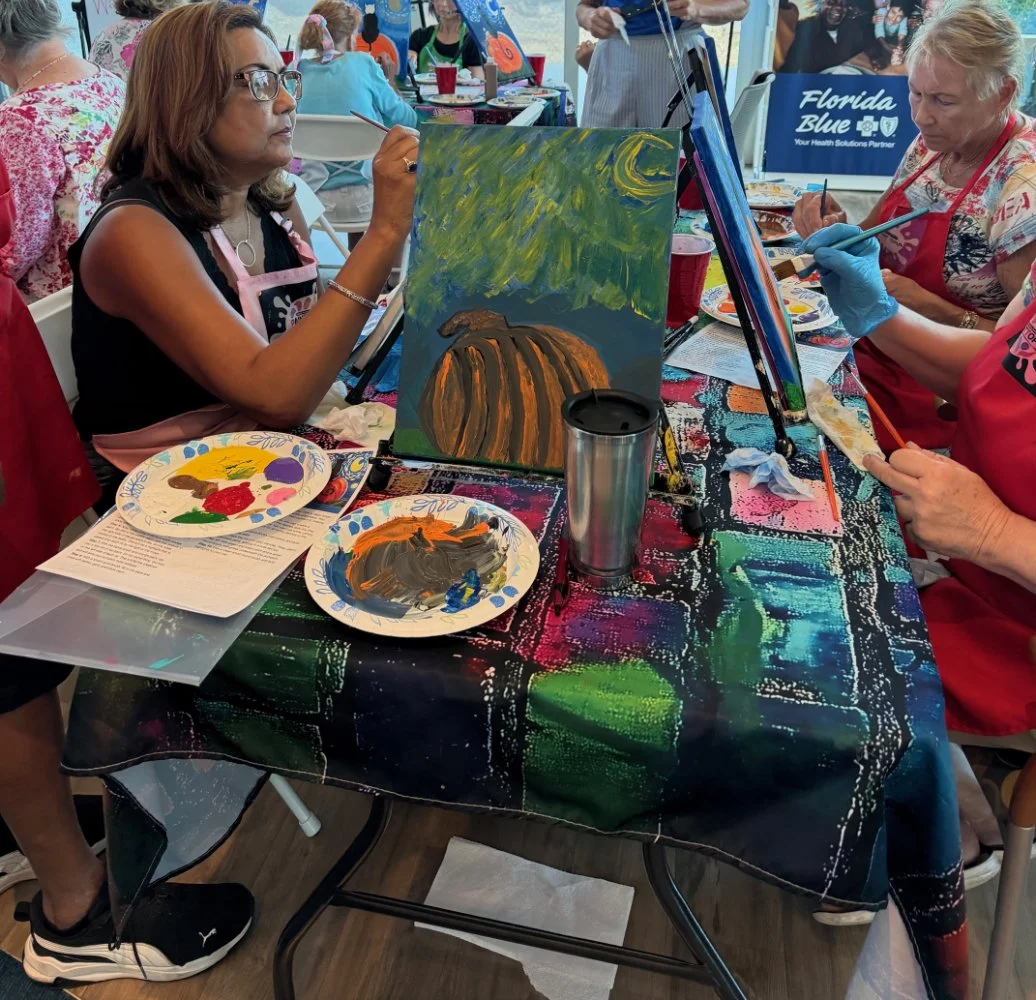 Women painting at a guided paint and sip event, creating a landscape scene with a pumpkin, night sky, and moon, seated at a table with colorful tablecloth and painting supplies.