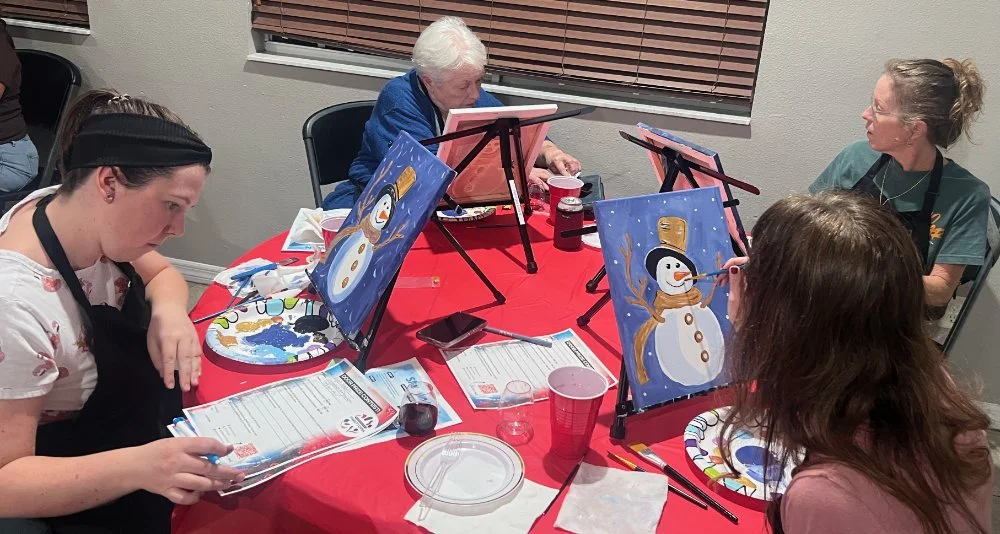 People painting snowman holiday pictures during a group event at a table covered with red tablecloth.
