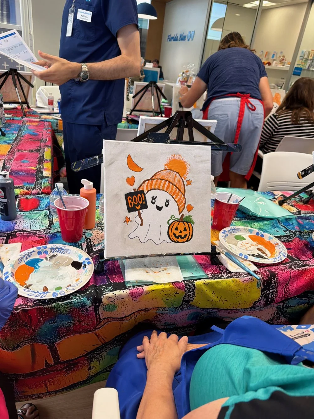 A lively art studio displaying a Halloween-themed painting of a cheerful ghost in a pumpkin hat, with fall decorations and painting supplies on a colorful, patterned table.