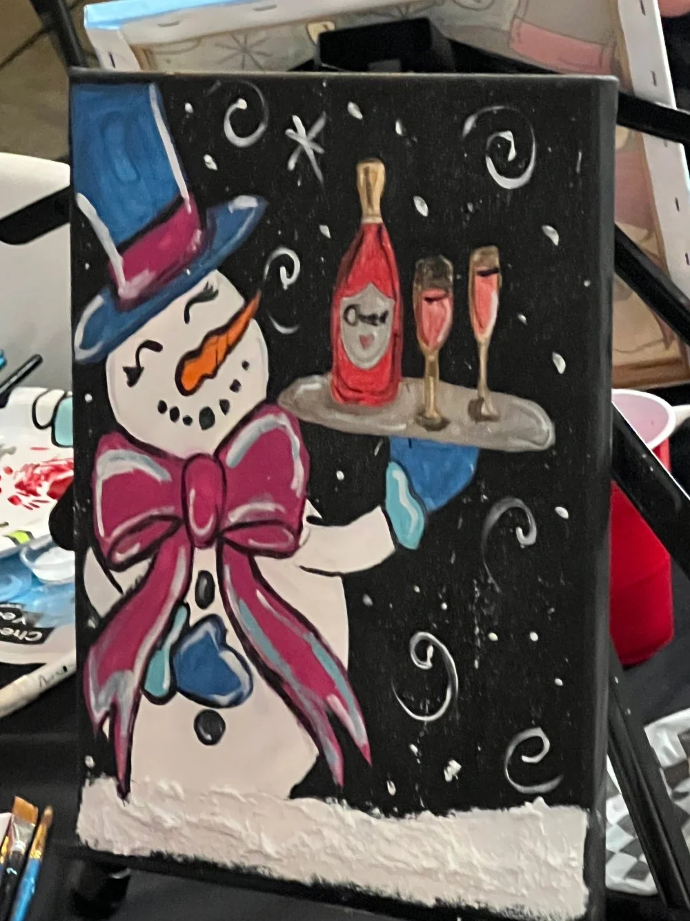 Colorful painting of a snowman with a pink bow tie, wearing a blue hat with pink band, holding a tray with a bottle and two glasses. The background is black with white swirl and dot decorations.