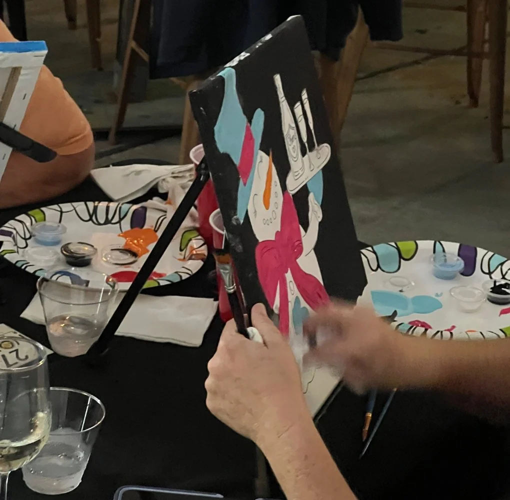 Person painting a colorful picture of a face with a red scarf on a black canvas in a social setting, with art supplies and drinks on the table.