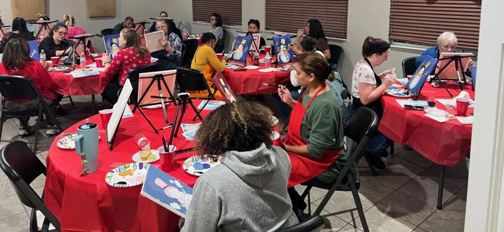 People participating in a group painting party with canvases, paints, and brushes at decorated tables.