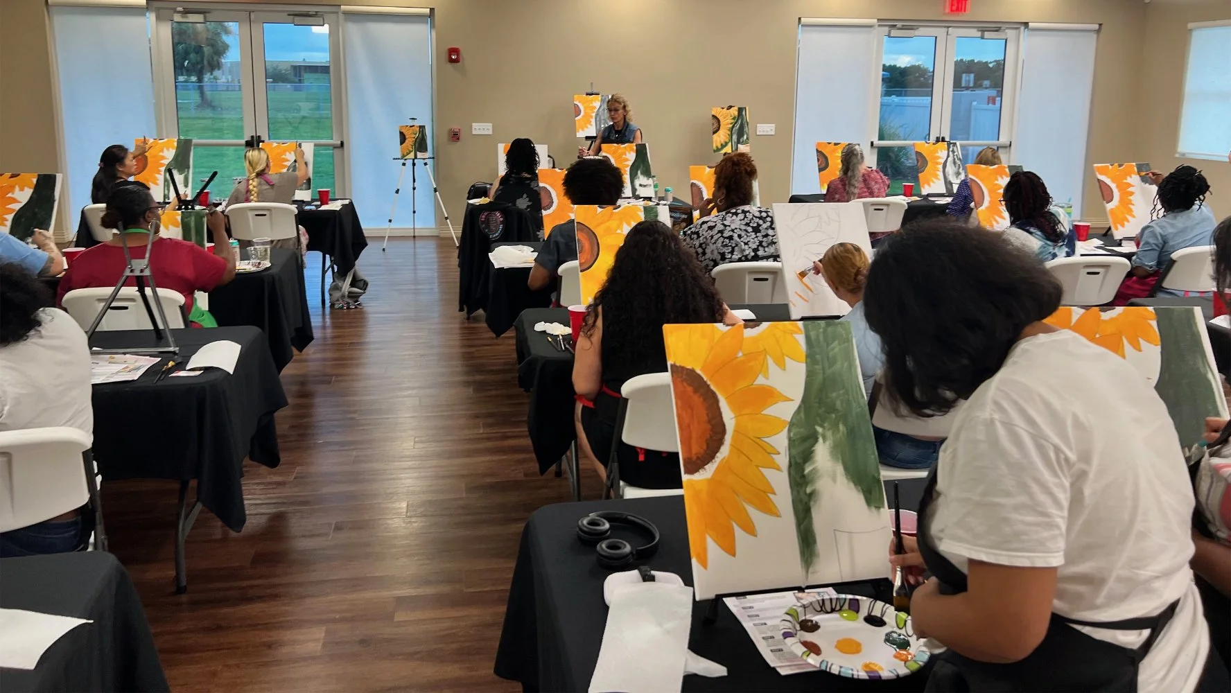 A group of people participating in a painting class, painting sunflowers on canvases while seated at black-covered tables in a bright room with large windows.