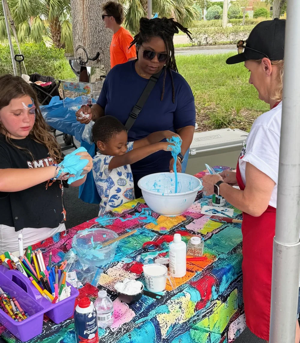 Children and adults making slime at a colorful outdoor table with art supplies, under a canopy with trees in the background.