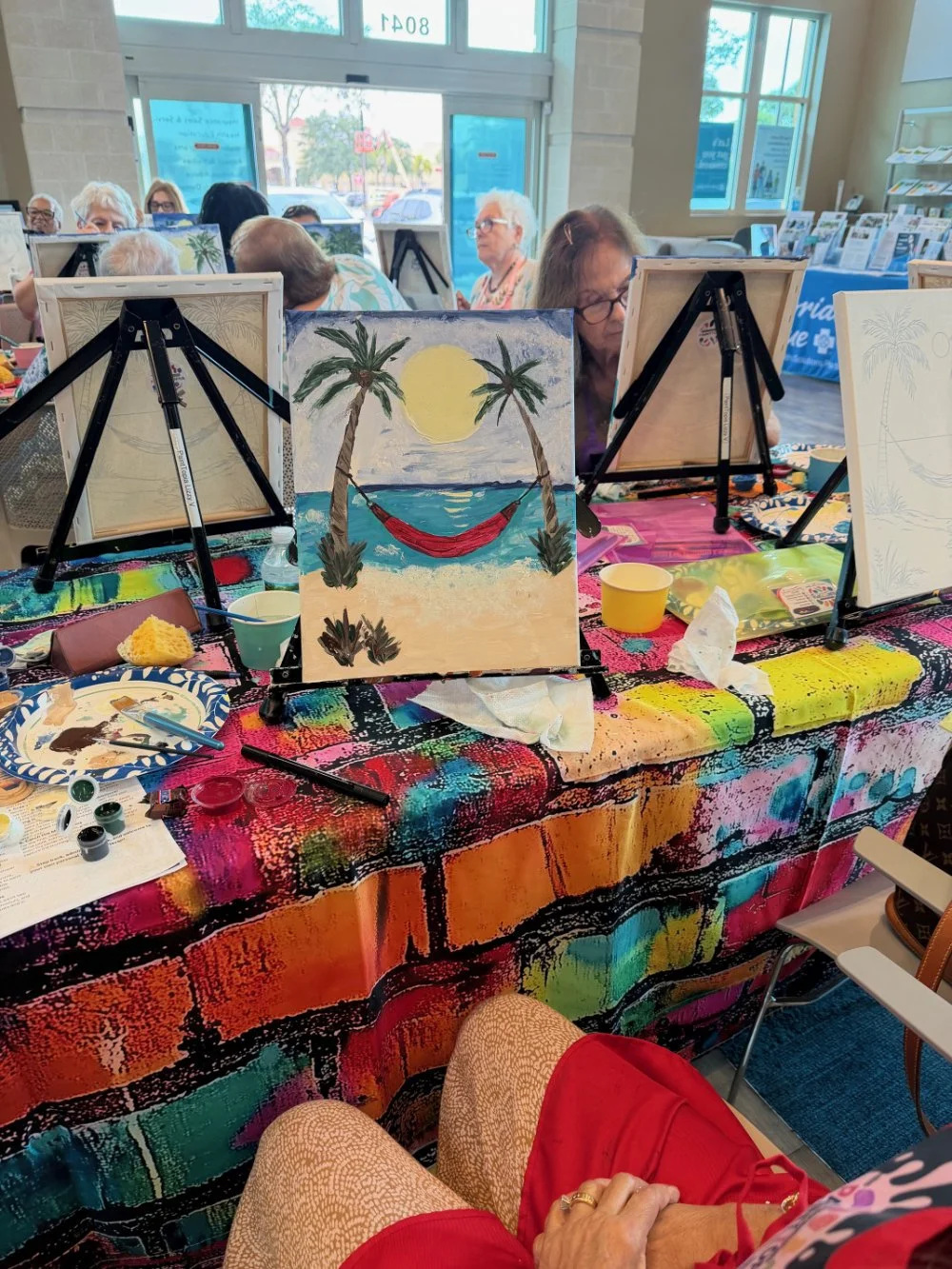 Group of older women painting tropical beach scenes with palm trees, a full moon, and a hammock on canvases during a painting class inside a bright room with large windows.