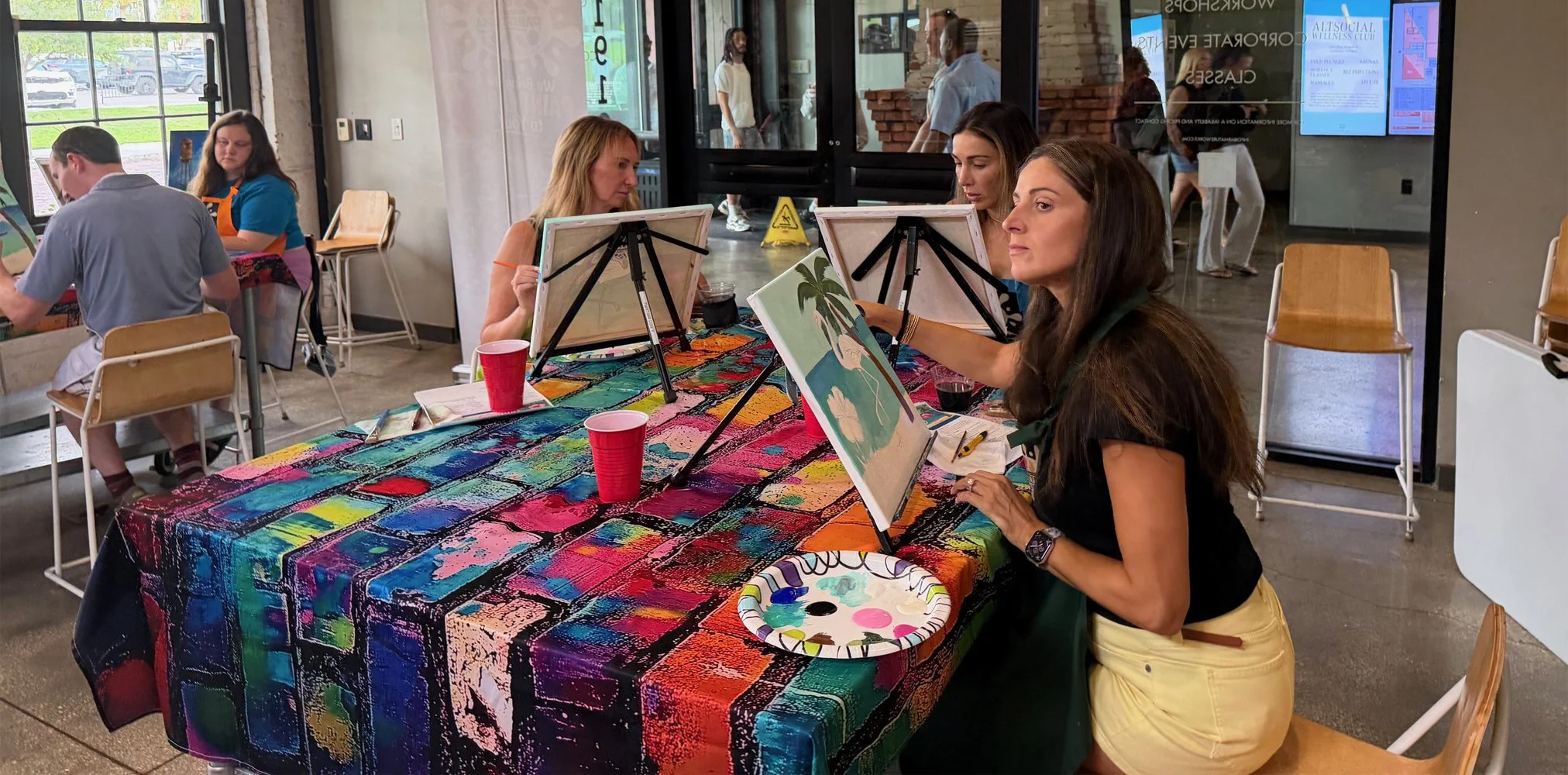 Women painting on canvases at a colorful table during an art class inside a cafe or community center.