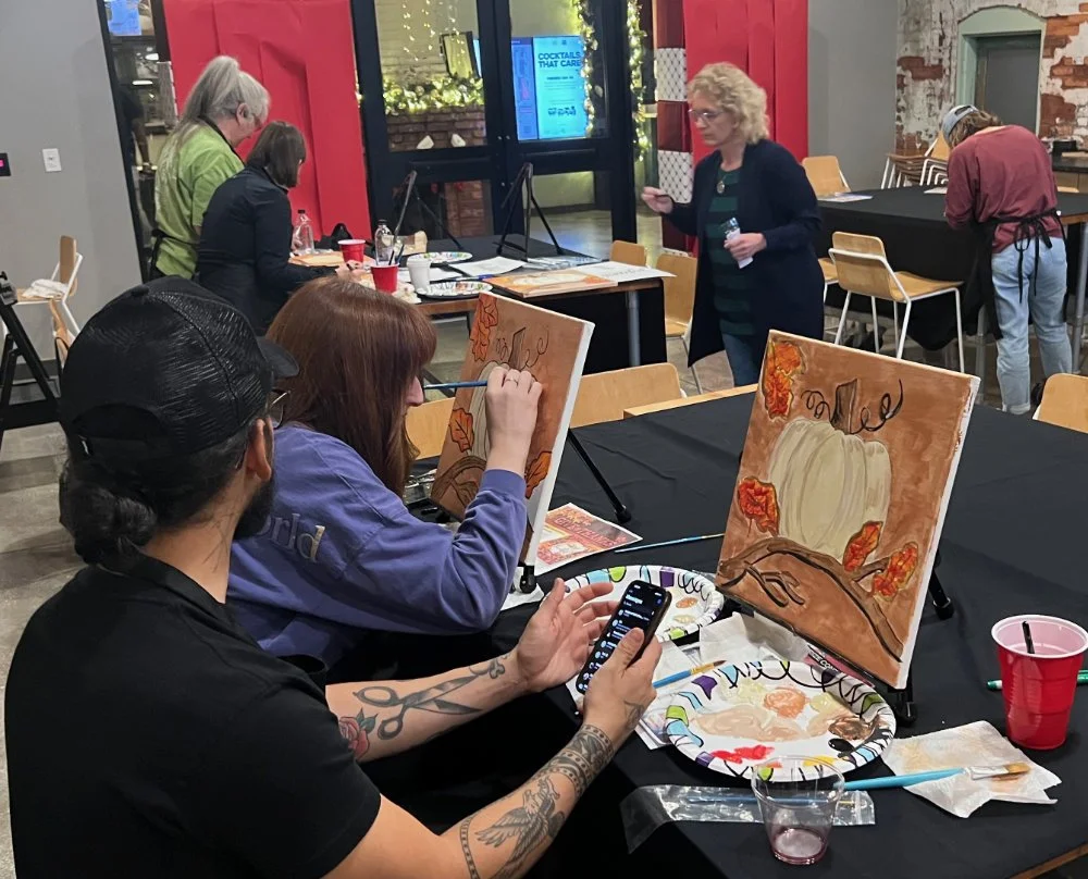 Women participating in a painting class, painting autumn-themed images with pumpkins and leaves on canvases at a table.