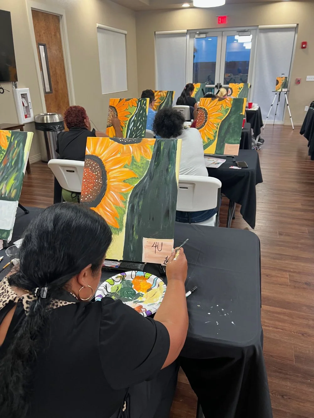 People participating in a paint and sip class, painting sunflower artwork on canvases at tables in a room with wooden floors and double glass doors.