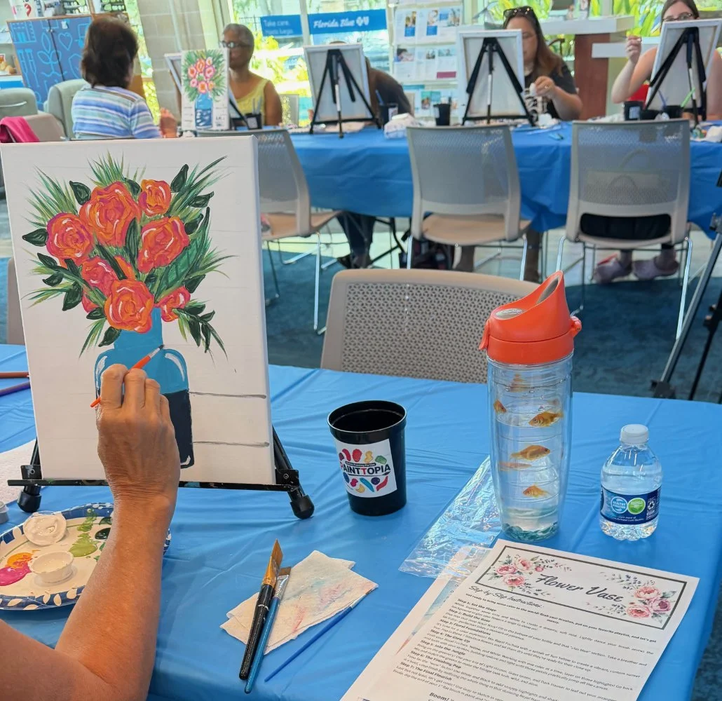 People painting floral arrangements on canvases at a craft event with blue tablecloths, paint supplies, and a photo of a flower vase with orange flowers.