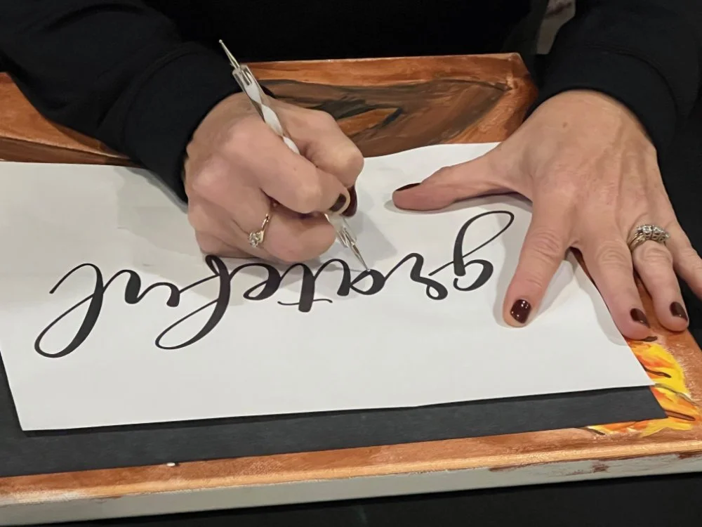 A person with manicured nails and rings is writing on a white piece of paper with a black marker, signing their name that reads 'Sealed'. The paper is on a wooden surface.