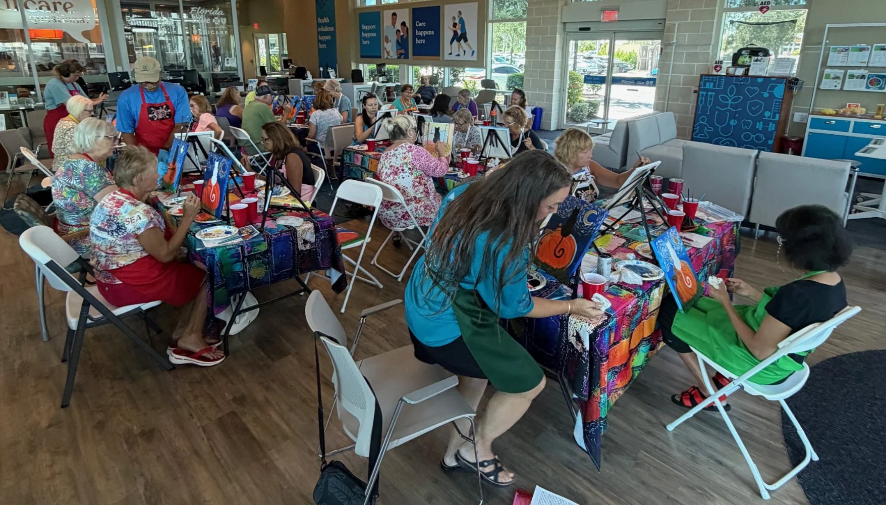 A group of people, mostly older women, are engaged in a painting class at a community center or store, with colorful paintings of pumpkins and a cat on the easels, and tables covered with vibrant tablecloths.