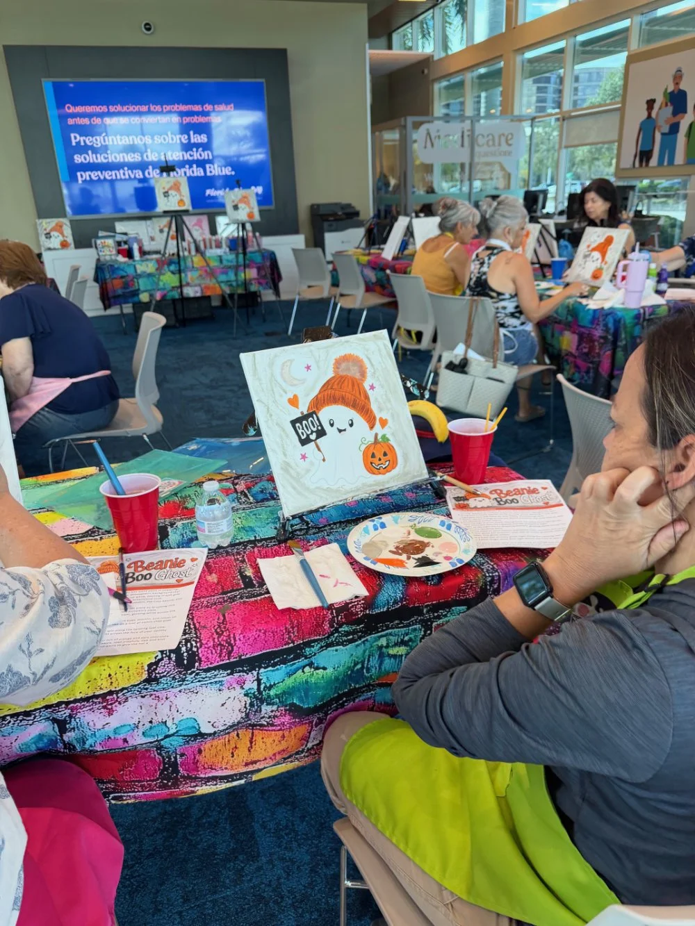 People participating in a Halloween-themed painting activity at an indoor event with colorful tables and a large screen displaying event information in the background.