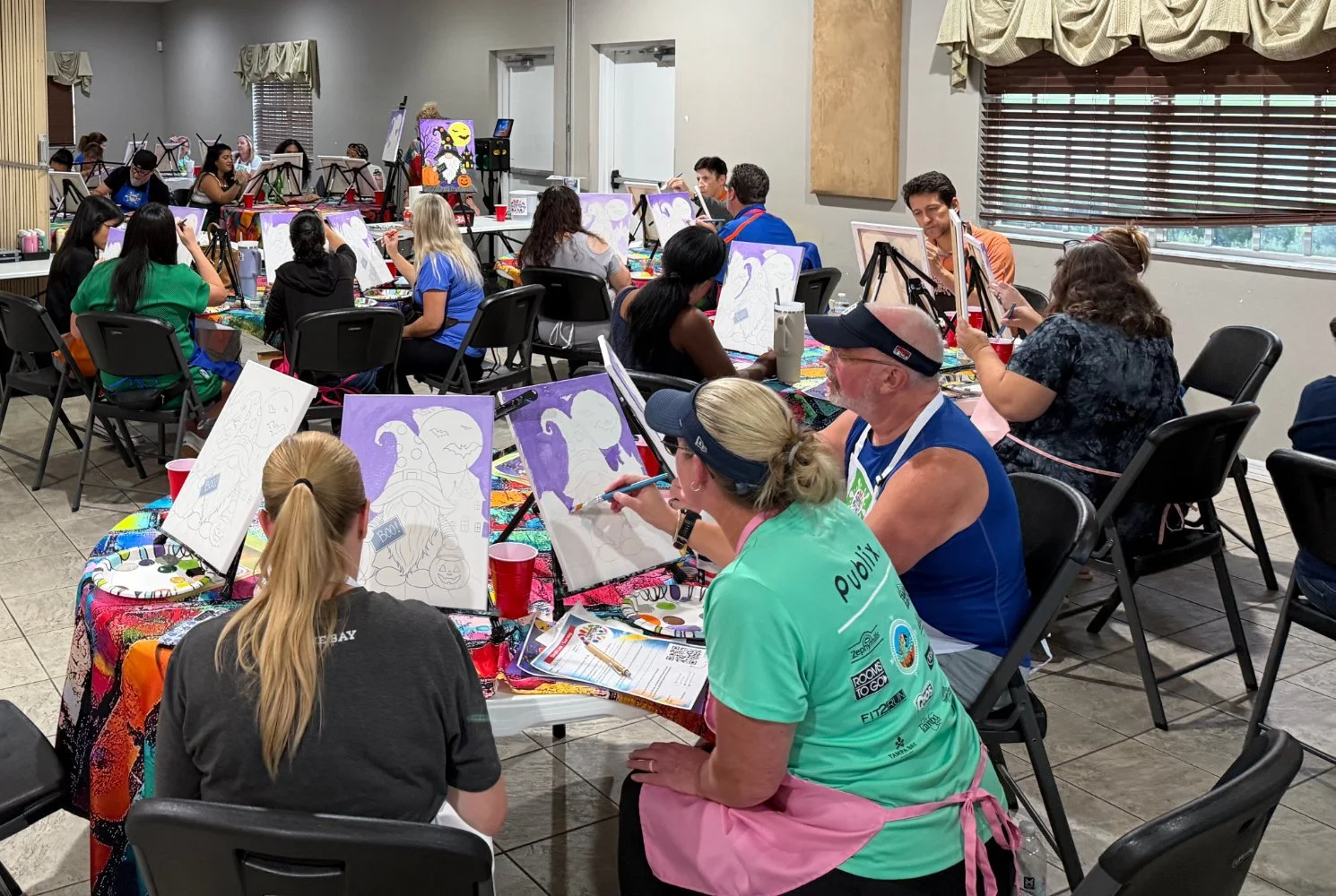 People participating in a group painting class, sitting at tables with canvases, paint supplies, and drinks, engaged in creating artwork in a well-lit room with windows and blinds.