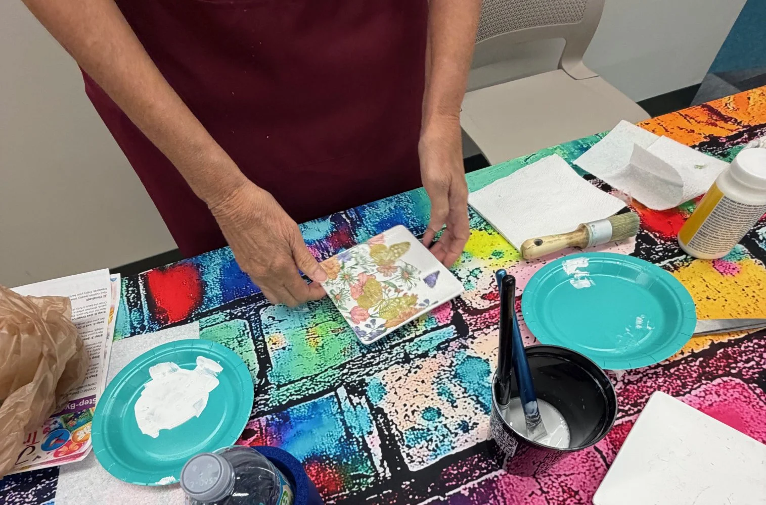 Person holding a decorative plate with floral design at colorful table with art supplies and paper plates.