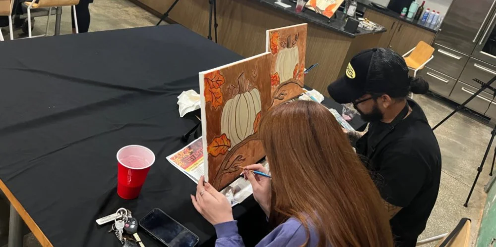 Two people are painting fall-themed scenes with pumpkins and leaves on canvases at a table in a room with kitchen cabinets and a red plastic cup.