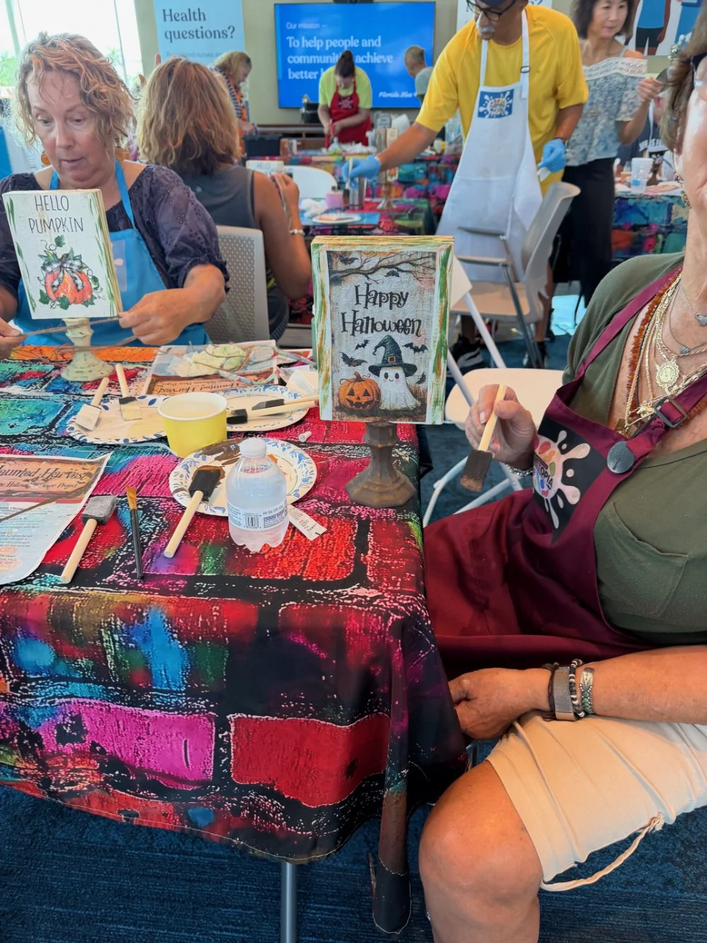 People participating in a DIY painting event at a table covered with colorful tablecloths. The table has art supplies, a water bottle, and small canvases. Two decorative signs on the table display Halloween themes, one reading 'Hello Pumpkin' with a 
