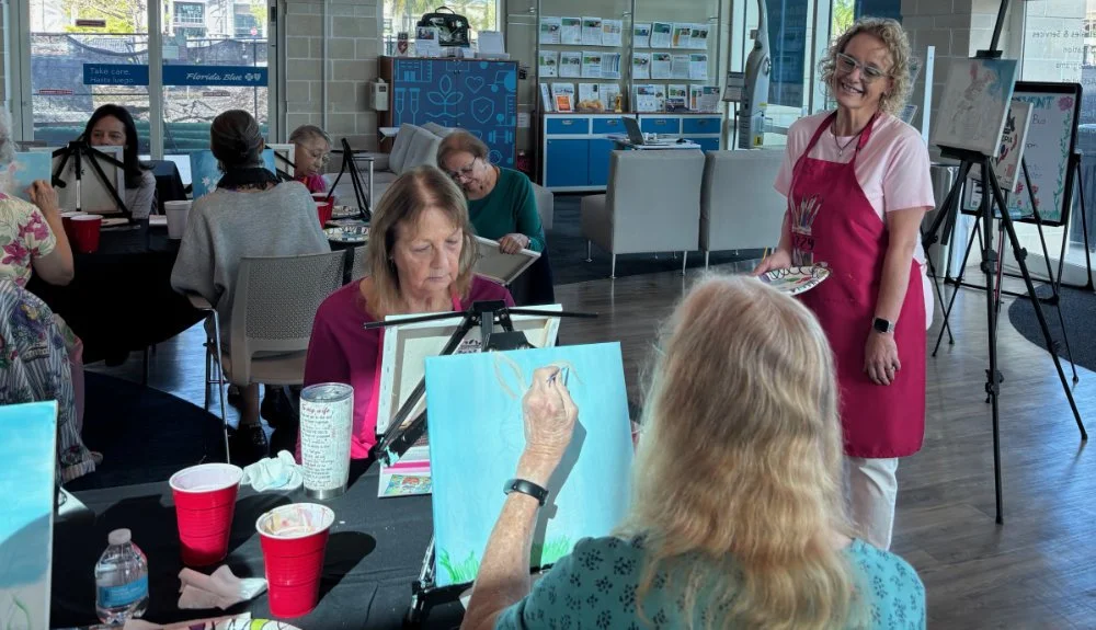 A group of older women participating in a painting class at an indoor location, with two women in the foreground working on their art and a smiling instructor wearing a pink apron holding a palette, overseeing the session.