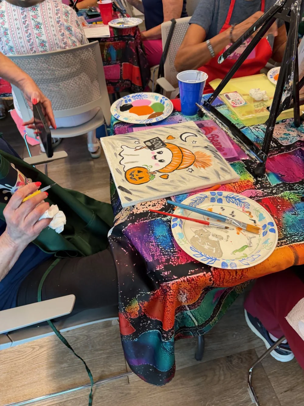 A table with a colorful, splattered paint-colored tablecloth featuring a Halloween-themed artwork of a cute ghost with a pumpkin, surrounded by art supplies like paintbrushes, paint, and plates, with people engaging in painting activity.