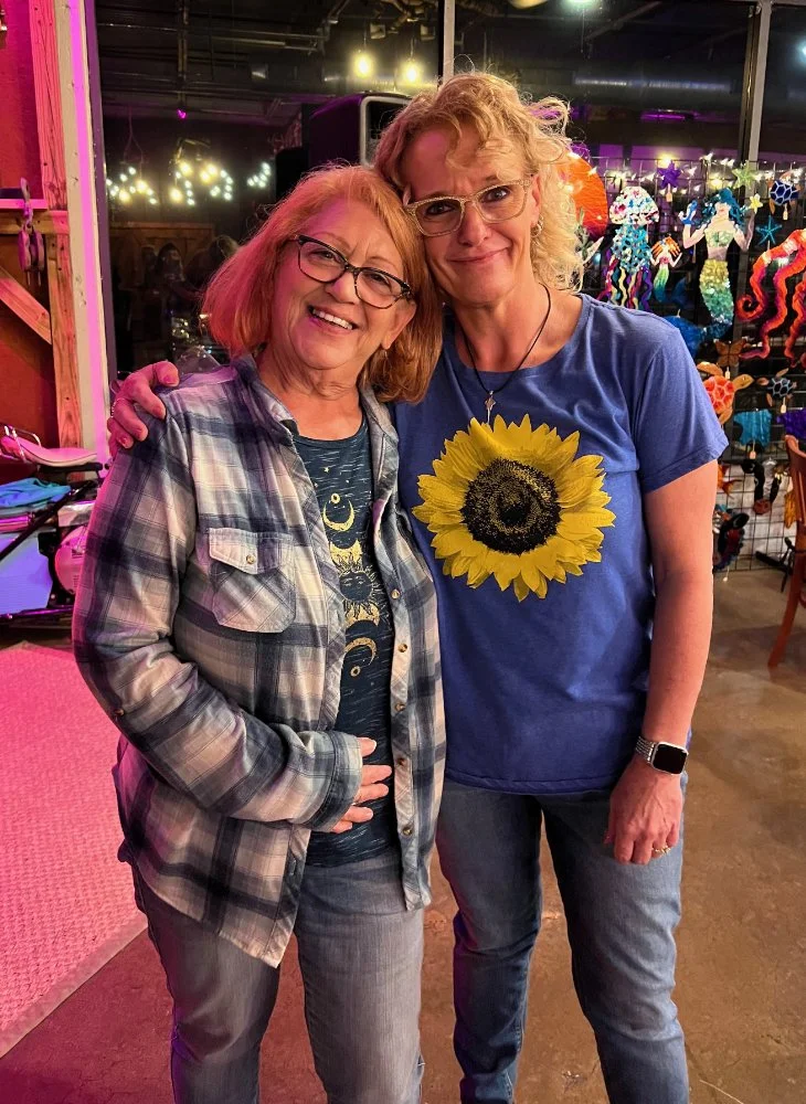 Two women standing close together, smiling at the camera, in a colorful, festive indoor setting with decorations and lights in the background.